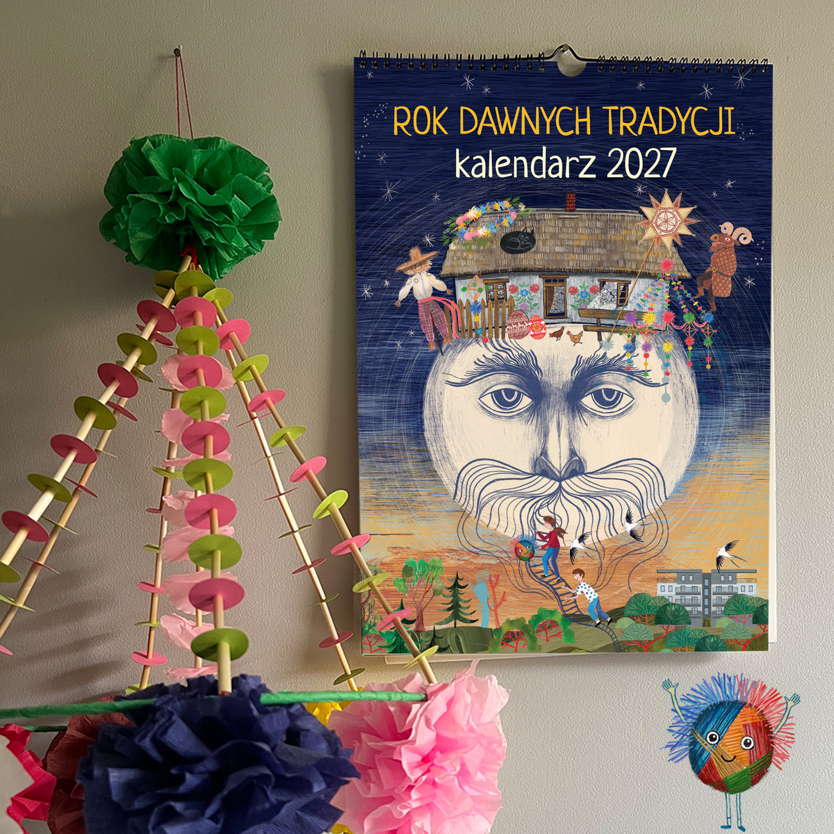 Granatowa okładka kalendarza „Rok Dawnych Tradycji 2027” z ilustracją ludowej postaci i chaty, zawieszona obok kolorowego pająka.