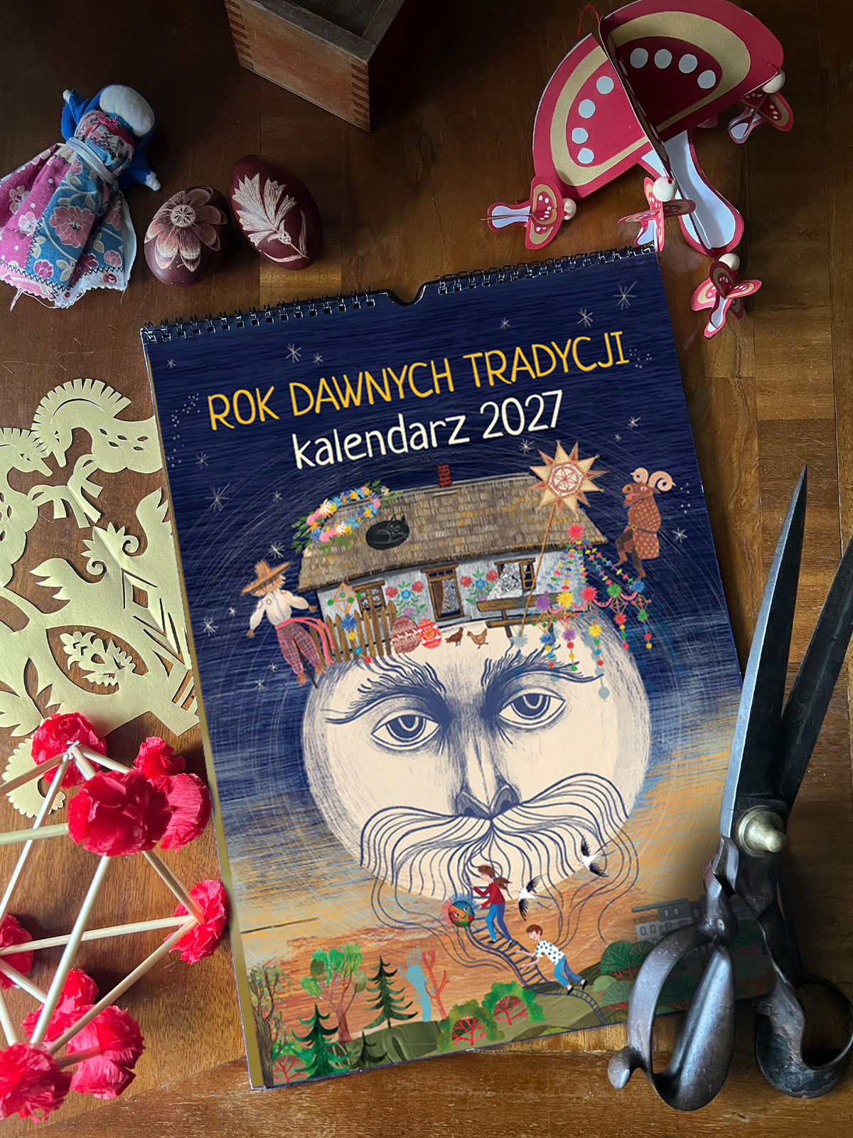 Granatowa okładka kalendarza „Rok Dawnych Tradycji 2027” z ilustracją ludowej postaci i chaty, zawieszona obok kolorowego pająka.