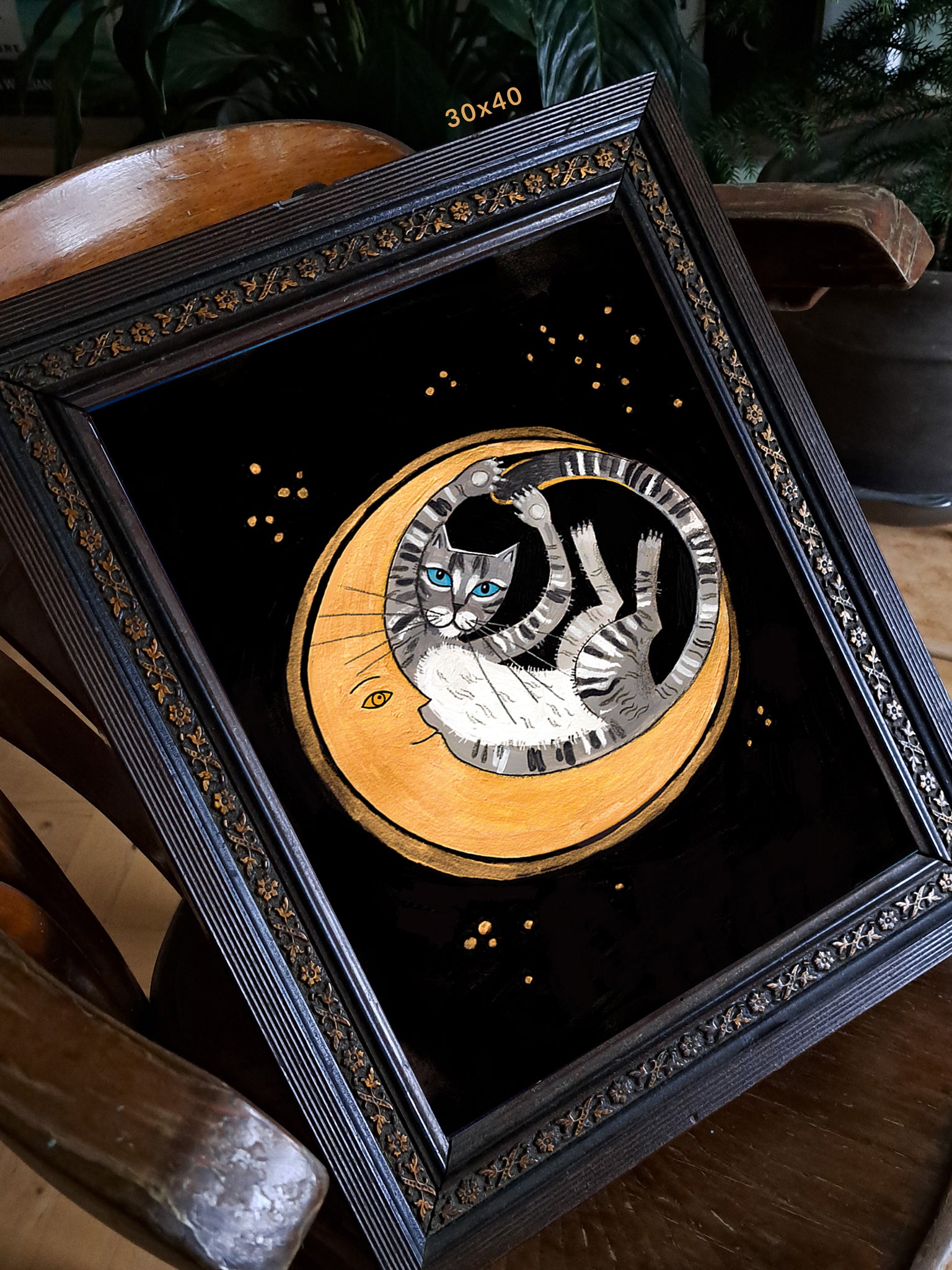 Wydruk „Pełnia Szczęścia" w rozmiarze 30x40 cm w czarnej zdobionej ramie na drewnianym krześle – ilustracja z kotem i księżycem jako dekoracja wnętrza